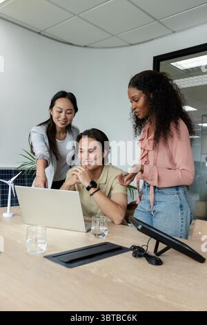 Diverse coworkers collaborating around laptop in meeting room with wind turbine and solar panel Stock Photo