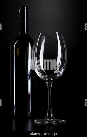 empty wine glass stands beside wine bottle against a deep black background Stock Photo
