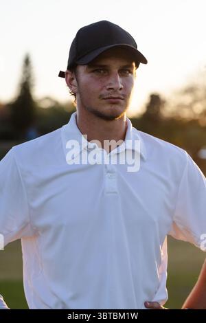Man standing on grassy sports field during golden hour wearing black baseball cap, white polo shirt Stock Photo