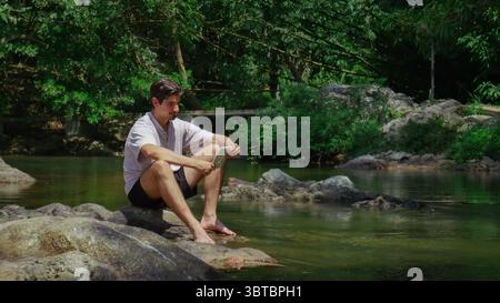 Young man sitting barefoot on rocks by a river in the jungle, reading a tablet, tropical forest scenery Stock Photo