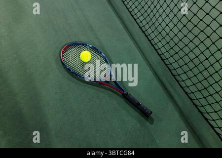 Tennis racket lying on green hardcourt with yellow ball resting on strings near black mesh net Stock Photo