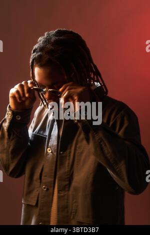 Young adult Black man adjusting sunglasses while standing against plain red background, head slightly tilted downward, wearing casual jacket, dreadlocks partially covering face Stock Photo