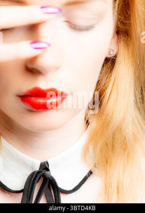 A close-up shot of a woman with red lips, closed eyes, and a hand covering her face. She has long, blonde hair and is wearing a white collared shirt w Stock Photo