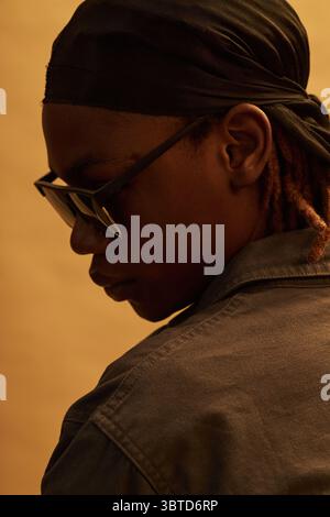 Black teenager wearing sunglasses and headscarf turning head away from camera, showing side profile and contemplative expression against plain background Stock Photo