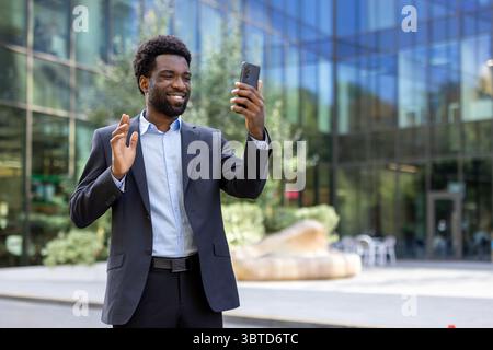 An African American businessman waves while video calling on his smartphone outside a modern office building, smiling and greeting someone on the call. Stock Photo