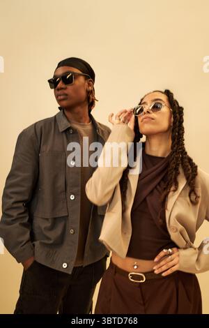 Young Black man and young Black woman wearing sunglasses posing confidently against plain background, woman adjusting sunglasses while both looking away from camera, stylish attitude Stock Photo
