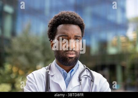 A serious-looking male doctor stands confidently, a stethoscope around his neck, outside of a modern building. Stock Photo
