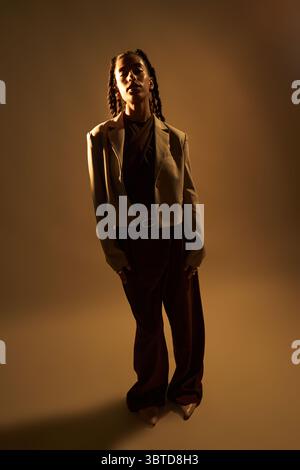 Portrait of young adult Black woman standing with relaxed posture, looking directly at camera, wearing business attire, illuminated by dramatic studio lighting, isolated on plain background Stock Photo