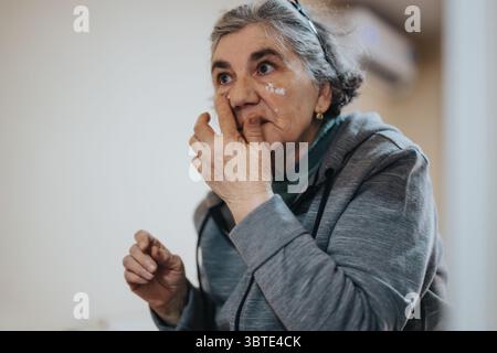 Elderly woman in casual attire thoughtfully touching her face indoors Stock Photo