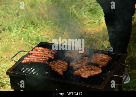 Barbecue scene with grilled sausages and pork steaks over hot coals, with a person standing near the grill in a grassy outdoor setting Stock Photo