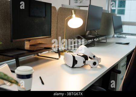 VR headset resting on desk under gold lamp casting warm light with monitors and coffee cup Stock Photo