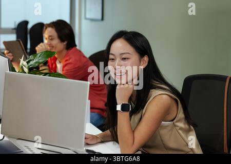 Asian coworkers collaborating using laptop and tablet at office desks with potted plant, copy space Stock Photo