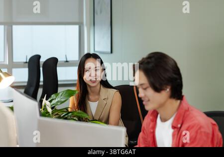 Asian coworkers smiling while working at office workstation using computer monitors with plant Stock Photo