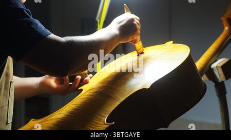 Luthier working on a cello in his workshop, applying varnish on the body of the instrument with a small brush, using a lamp to better see details and Stock Photo
