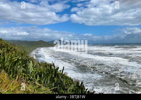 Dramatic coastal landscape on New Zealand’s South Island West Coast with waves, dark sand beach and distant hills under a dynamic sky Stock Photo