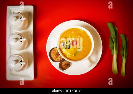 A vibrant bowl of creamy yellow curry noodles garnished with chili, mushrooms, and greens. Set against a bold red background. Stock Photo