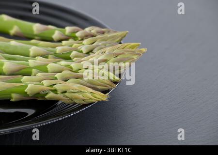 Close-up shot of fresh asparagus spears arranged on a black plate, set against a dark gray background, suitable for culinary or healthy eating themes. Stock Photo