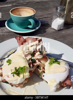 Eggs Benedict with crispy bacon, poached eggs, and hollandaise sauce served on toast, with coffee on a wooden table in natural light Stock Photo