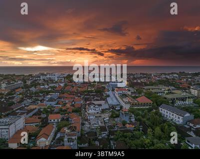 Aerial view of terracotta rooftops nestled between lush trees, meeting the ocean under a fiery sunset sky, Badung, Bali, Indonesia. Stock Photo