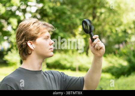 Handsome young caucasian man listen music and hand electric fan suffering from heat at park. Overheated man suffering from heat attack uses an electri Stock Photo