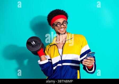 Young afro haired man wearing retro sportswear smiling with weight in hand and using smartphone Stock Photo