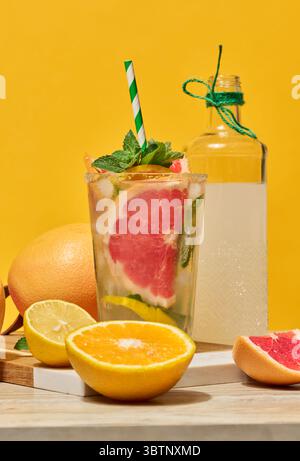 A clear glass with a cold drink, grapefruit slices, ice and mint, with a green striped straw, on a yellow background. Sliced ​​citrus fruits lie nearb Stock Photo