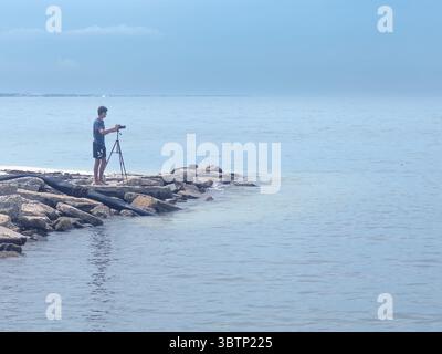 **Caption:** Man with a tripod-mounted camera stands on the rocky jetty, near the sandy shore of Mexico Beach, capturing the tranquil Gulf of Mexico w Stock Photo