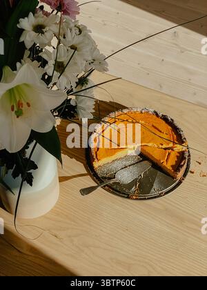 From above you can see a pumpkin pie cut open next to a beautiful bouquet of flowers in a vase on an ash wood table standing on a glass floor. Stock Photo