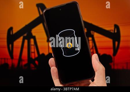BOGOTA, COLOMBIA - JULY 14 OF 2025 A male hand holding a smartphone with an oil barrel logo and a blurred oil platform in dusk at background Stock Photo