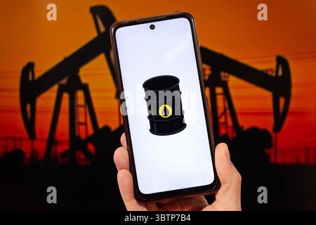BOGOTA, COLOMBIA - JULY 14 OF 2025 A male hand holding a smartphone with an oil barrel logo and a blurred oil derrick silhouette in sunset at backgrou Stock Photo