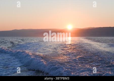 Calm sea at sunset with soft waves and distant hills, glowing in warm tones of orange and purple under a clear sky. Stock Photo