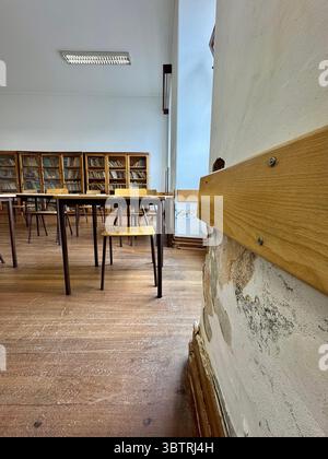 Old classroom with wooden desks and visible wall damage, school interior Stock Photo