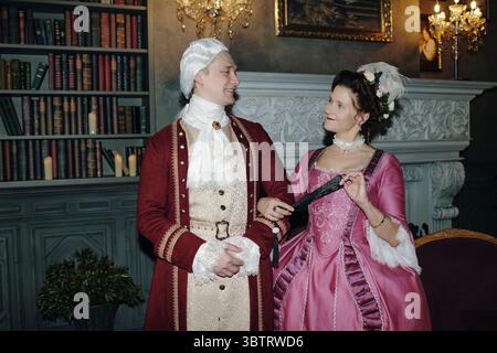 Caucasian middle aged man wearing powdered wig and ornate suit standing beside Caucasian middle aged woman in elaborate pink Baroque dress holding black fan, both smiling indoors Stock Photo