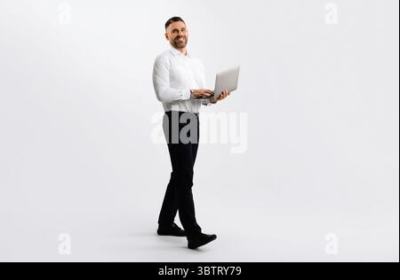 Full length shot of businessman with laptop walking over light grey studio background, male entrepreneur using computer Stock Photo