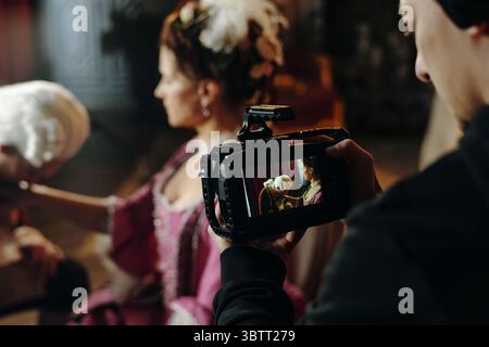 Caucasian young adult woman wearing Baroque style costume sitting while being filmed by young adult Caucasian man holding professional video camera during historical reenactment Stock Photo