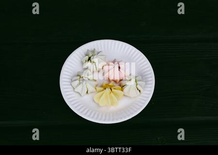Colorful meringues on a white plate, a delectable dessert. A delightful plate of multicoloured meringues sits on a dark brown surface, a sweet treat f Stock Photo