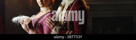 Caucasian young adult woman and Caucasian young adult man wearing Baroque period costumes standing together in historical setting, woman holding feathered fan, both facing camera Stock Photo