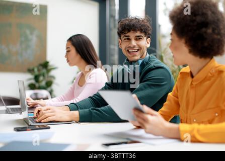 Happy diverse students sitting in classroom and communicating, using gadgets while being absorbed in studying Stock Photo