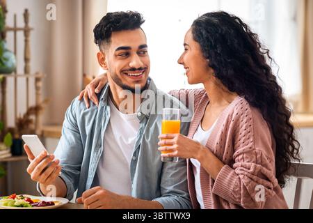 Happy Young Arab Couple Using Smartphone During Breakfast In Kitchen Stock Photo