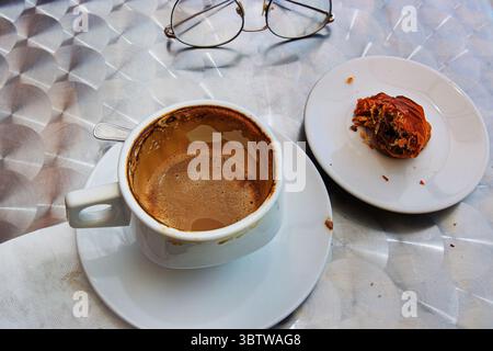 The white ceramic cup with  cappuccino leftovers with creamy foam, last bite of sfogliatella paistry and glasses on a metallic table in outdoor cafe. Stock Photo
