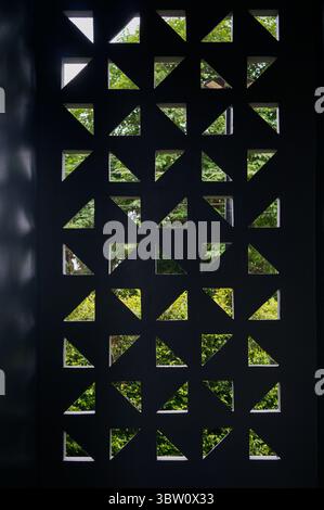 Geometric pattern of triangular and square cutouts in a dark panel with green foliage visible through the openings. Stock Photo