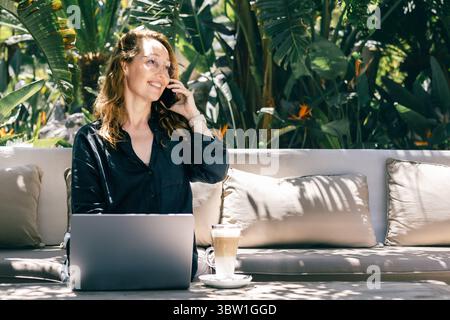 Smiling Woman Talking on Phone While Working on Laptop Outdoors in Tropical Garden with Coffee Stock Photo