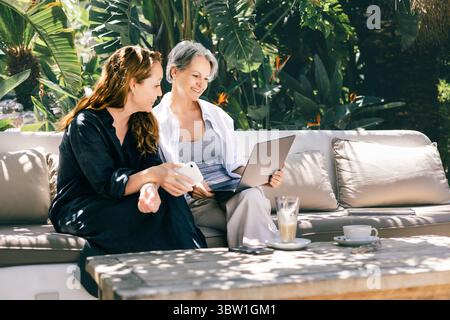 Happy mature women working remotely together with laptop and phone in tropical garden setting Stock Photo