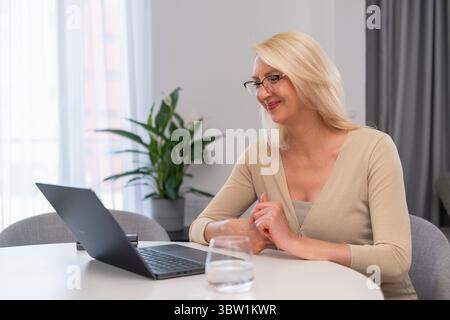 Middle aged blonde psychologist talking to patient online via laptop. Mature woman therapist in beige outfit having video call consultation. Concept of modern remote therapy and mental wellness. Stock Photo