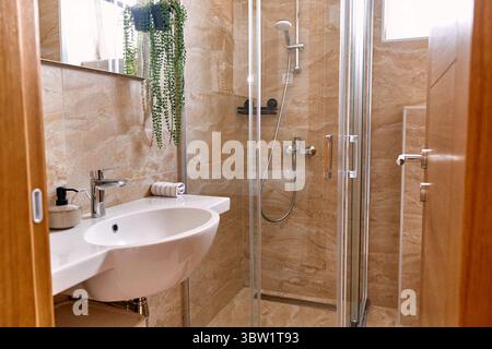 A modern bathroom featuring beige tiles, a glass shower enclosure, and a floating sink, creating a natural and elegant ambiance. contains a small plan Stock Photo