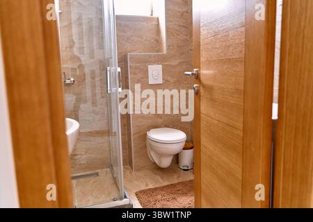 A modern bathroom with wooden doors, featuring stone wall tiles, a wall-mounted toilet, and a glass shower enclosure, offering a compact yet stylish d Stock Photo