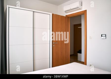 A minimalist bedroom interior showcasing a sleek wardrobe and wooden door, complemented by an air conditioning unit, emphasizing modern, clean design. Stock Photo