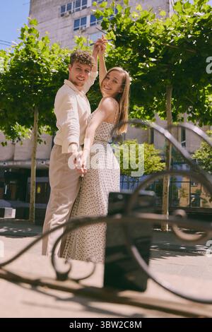 Happy young couple posing playfully for smartphone photo while holding hands on sunny city street Stock Photo