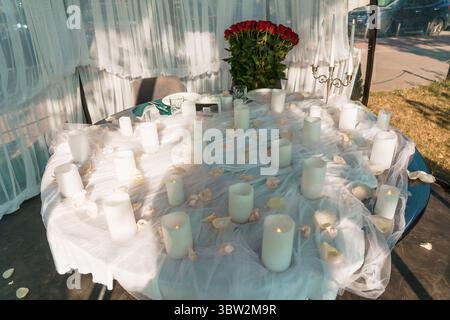 An intimate outdoor proposal scene features a table adorned with red roses, white candles, and tulle, suggesting a romantic gesture, private celebrati Stock Photo