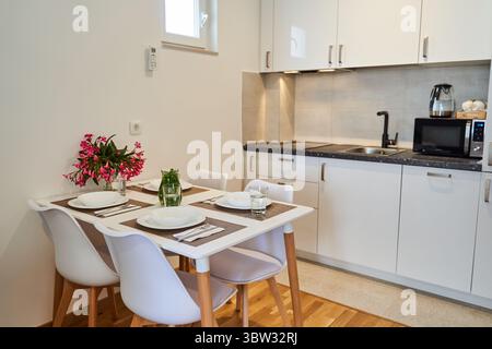 A modern kitchen showcasing a minimalist design with a dining table set for two, white cabinets, and a compact layout, creating a cozy and functional Stock Photo
